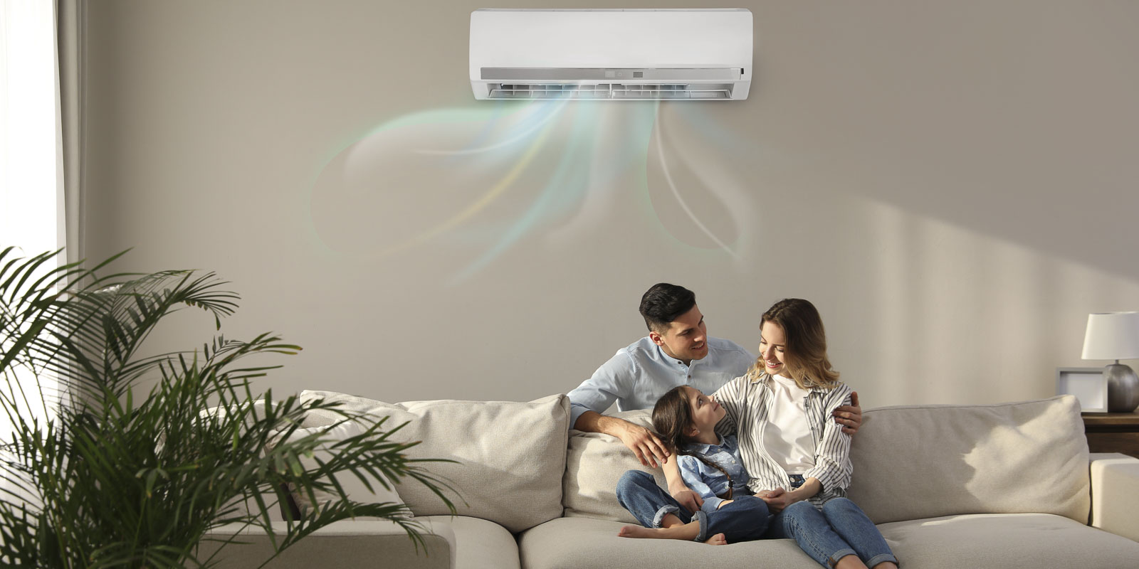 Im Wohnzimmer sitzt eine Frau mit Tochter auf dem Sofa und der Mann steht hinter den beiden. Alle schauen sich glücklich an. An der Wand im Hintergrund ist eine Klimaanlage, die Frische Luft in den Raum bringt.