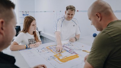 Eine Gruppe von vier Personen diskutiert über einen detaillierten Plan auf einem Tisch, während sie konzentriert schauen.