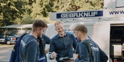 Im Hintergrund eines Lieferwagens stehen vier Männer, die sich angeregt unterhalten und dabei freundlich lachen.