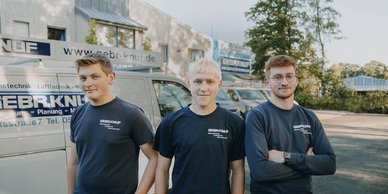 Die drei Männer tragen einheitliche T-Shirts mit dem Logo der Firma und posieren selbstbewusst vor einem Lieferwagen.