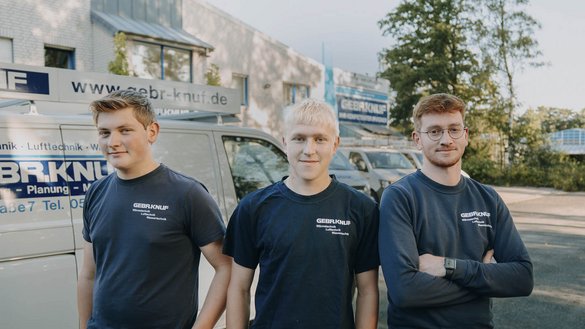 Die drei Männer tragen einheitliche T-Shirts mit dem Logo der Firma und posieren selbstbewusst vor einem Lieferwagen.