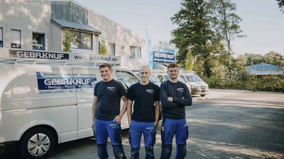 Drei Handwerker in blauen Arbeitsanzügen stehen lächelnd vor einem Firmenfahrzeug und einem modernen Gebäude im Hintergrund.