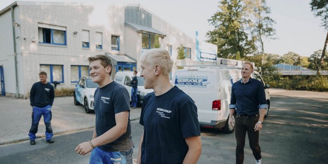 Eine Gruppe von vier Männern in Arbeitskleidung lacht und unterhält sich vor einem Firmengebäude mit blauen Fenstern.