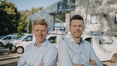 Die beiden Männer in hellblauen Hemden posieren selbstbewusst vor ihrem Geschäft, das im Hintergrund sichtbar ist.