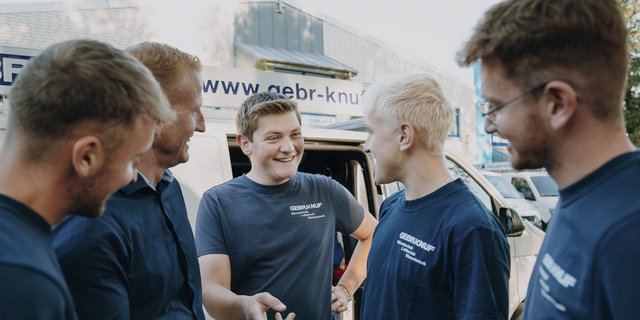 Eine Gruppe von fünf Männern lächelt und tauscht freundliche Worte aus, während sie vor einem Lieferwagen stehen.