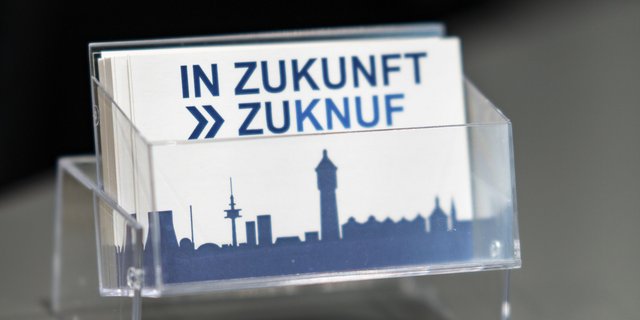 Eine transparente Halterung zeigt Karten mit dem Schriftzug 'IN ZUKUNFT' und einer stilisierten Stadtsilhouette im Hintergrund.