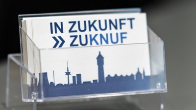 Eine transparente Halterung zeigt Karten mit dem Schriftzug 'IN ZUKUNFT' und einer stilisierten Stadtsilhouette im Hintergrund.