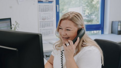 Eine lächelnde Frau spricht am Telefon, während sie an einem Schreibtisch mit einem Computer sitzt und einen hellen Raum umgibt.