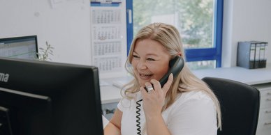 Eine lächelnde Frau spricht am Telefon, während sie an einem Schreibtisch mit einem Computer sitzt und einen hellen Raum umgibt.