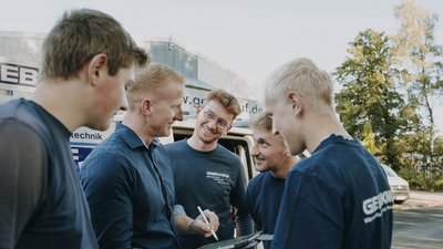 Eine Gruppe von fünf Männern lächelt und unterhält sich, während sie vor einem Lieferwagen mit dem Logo 'GEBR.KNUF' stehen.