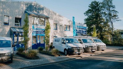Ein modernes Firmengebäude mit blauen Akzenten, umgeben von Bäumen und mehreren weißen Lieferwagen auf dem Parkplatz.