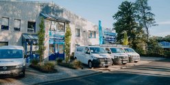Ein modernes Firmengebäude mit blauen Akzenten, umgeben von Bäumen und mehreren weißen Lieferwagen auf dem Parkplatz.