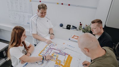 Vier Personen stehen um einen großen Plan, während sie angeregt über Details diskutieren und auf verschiedene Bereiche zeigen.