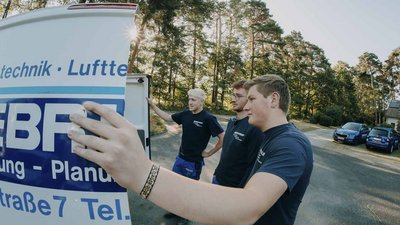 Drei junge Männer stehen vor einem Lieferwagen im Wald, während einer von ihnen ein Schild anbringt.
