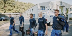 Eine Gruppe von Handwerkern verlässt ein Gebäude, jeder trägt Ausrüstung und zeigt eine freundliche, entspannte Stimmung.