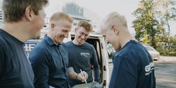 Die Gruppe von Männern diskutiert angeregt, während sie an einem Fahrzeug im Hintergrund stehen.
