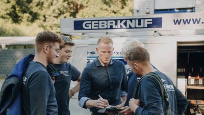 Eine Gruppe von fünf Männern in Arbeitskleidung diskutiert vor einem Lieferwagen, während einer Notizen macht.