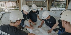 Sechs Bauarbeiter in Schutzhelmen studieren gemeinsam Baupläne auf einem Tisch in einer Baustelle mit Holzrahmen.