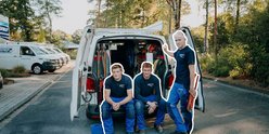 Drei Handwerker in blauen Arbeitsanzügen posieren vor einem Lieferwagen, umgeben von Bäumen und einem Parkplatz.