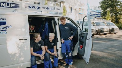Ein Lieferwagen mit dem Logo 'GEBR-KNUF' steht offen, während drei Männer in blauen Hosen miteinander lachen.