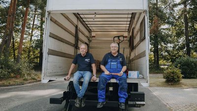 Die beiden Männer in Arbeitskleidung lächeln, während sie auf der Ladefläche eines Transporters sitzen, bereit für den Umzug.