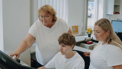Eine Frau zeigt einem jungen Mann an einem Computer etwas, während eine andere Frau interessiert zuschaut.